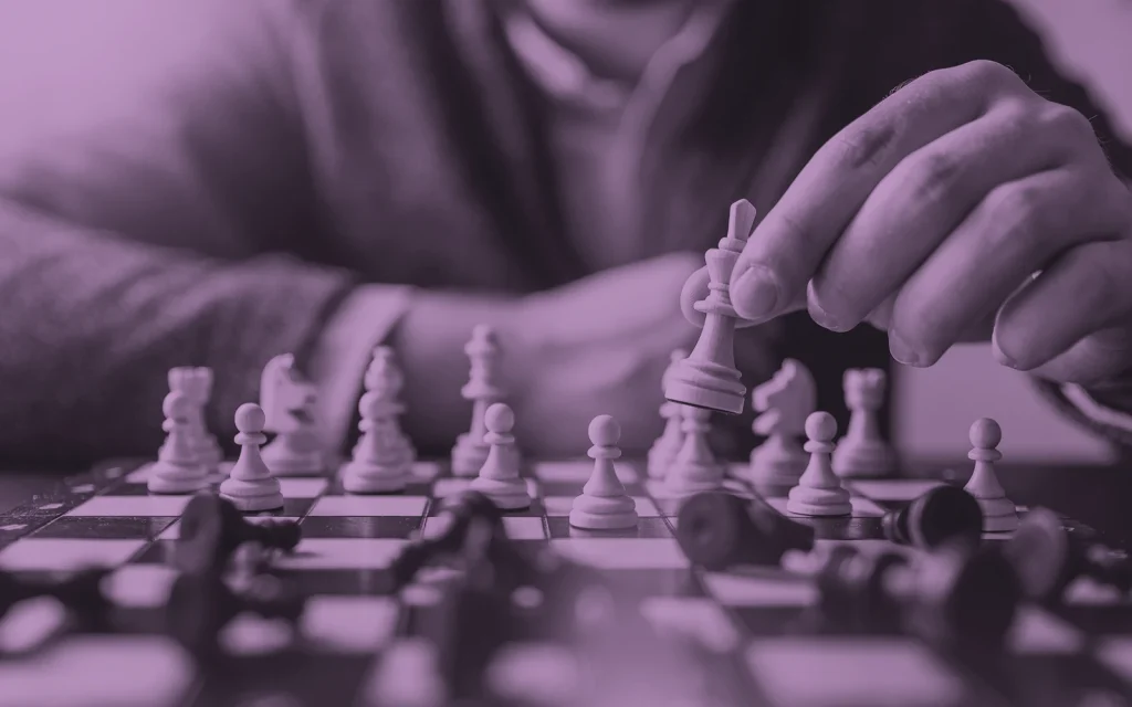 Purple-toned image of a person's hand carefully moving a white queen across a chess board mid-game, with fallen black pieces around it.
