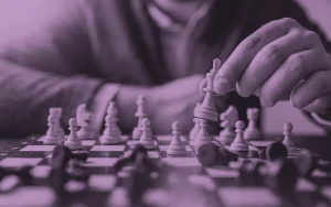 Purple-toned image of a person's hand carefully moving a white queen across a chess board mid-game, with fallen black pieces around it.