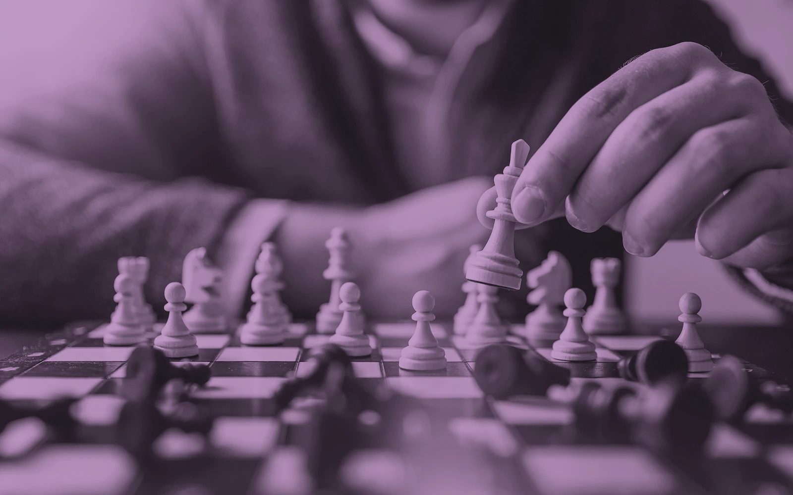 Purple-toned image of a person's hand carefully moving a white queen across a chess board mid-game, with fallen black pieces around it.