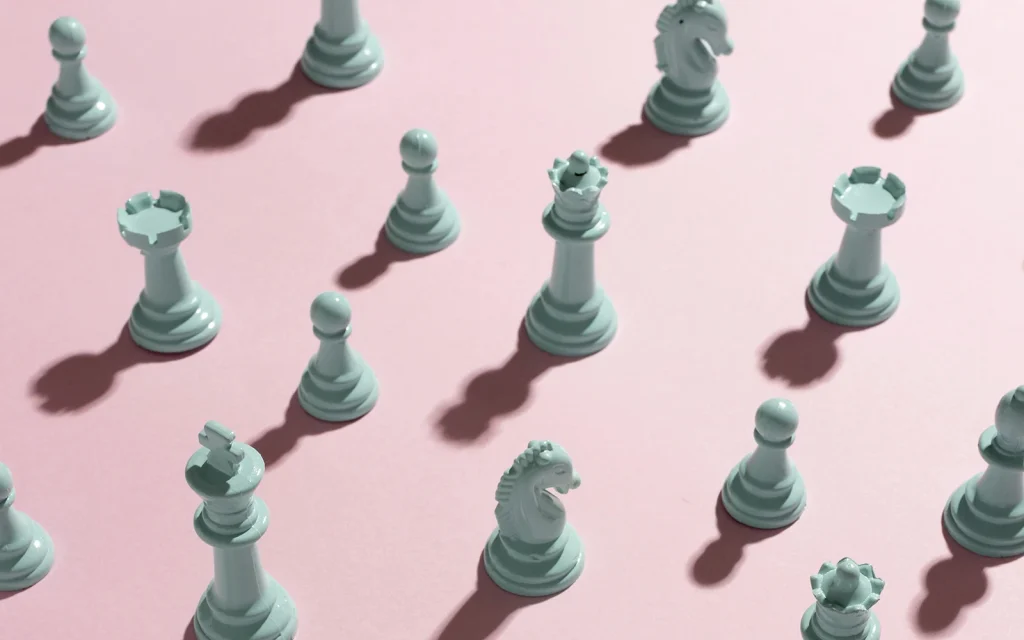 Overhead view of mint-green chess pieces arranged in a diagonal pattern on a pastel pink background, casting long shadows.