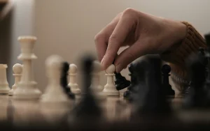 Close-up of a hand reaching down to move a white pawn on a chess board, with black and white pieces in soft focus.