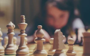 Warm-toned close-up of wooden chess pieces on a board with a child blurred in the background, focused on the game.