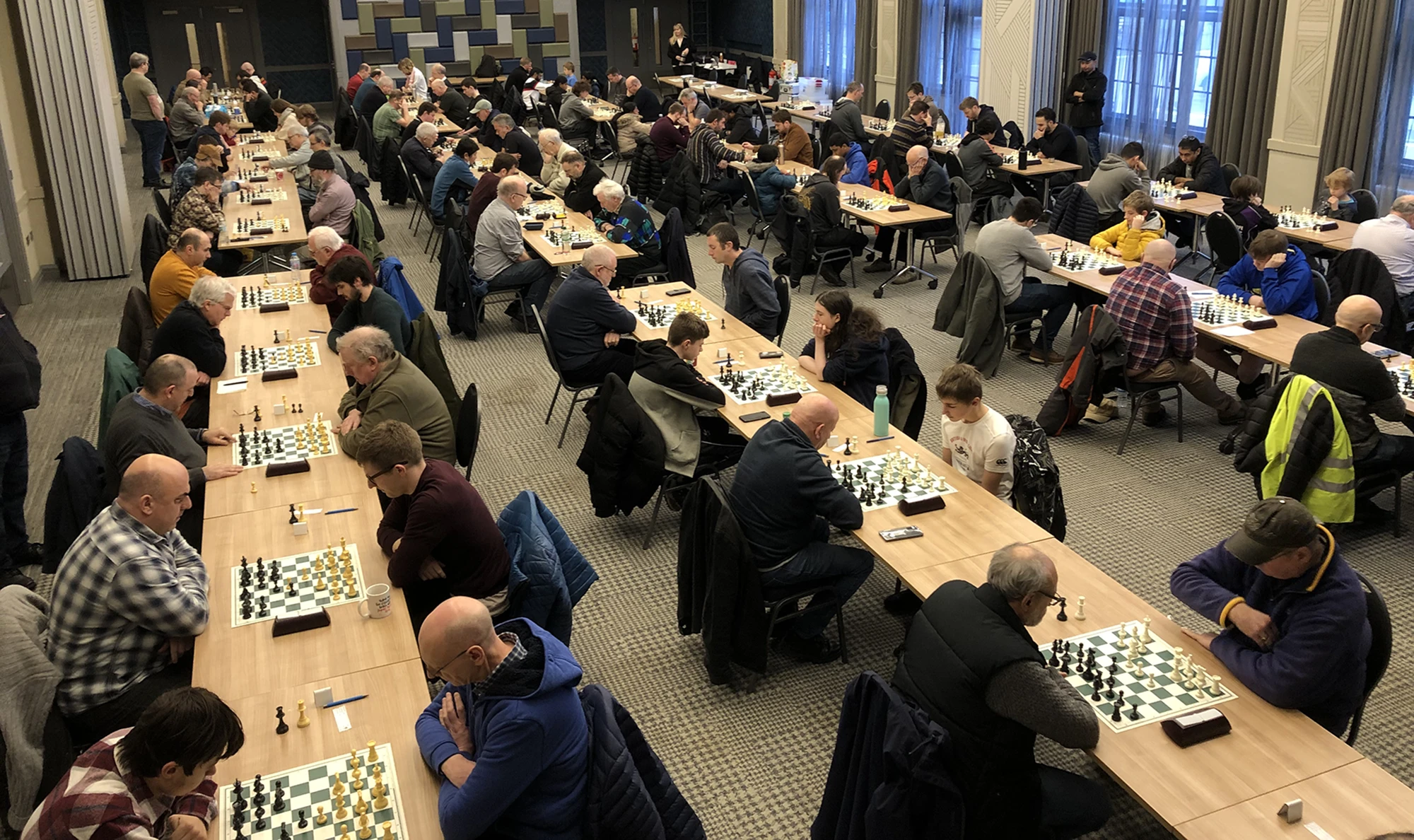 Wide view of a HERCA chess competition, with rows of players seated at long tables concentrating on their games in a large hall.