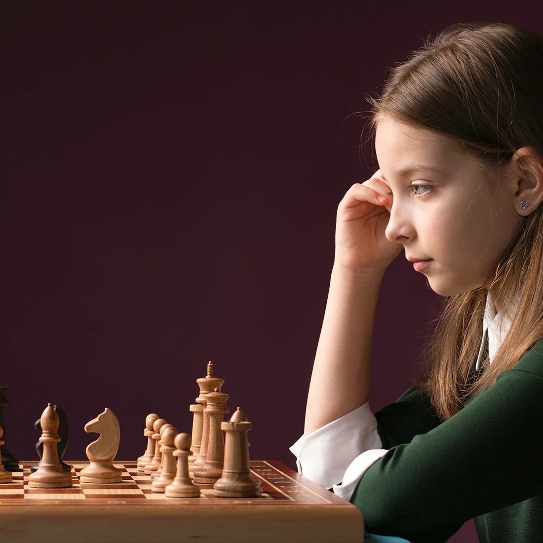 A young girl in a school uniform with green cardigan resting her chin on her hand, gazing thoughtfully at a wooden chess board against a deep burgundy background.
