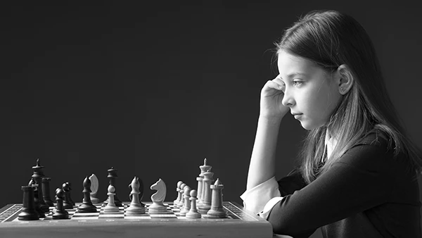 Black and white profile photograph of a young girl resting her chin on her hand, studying a chess board in front of her.